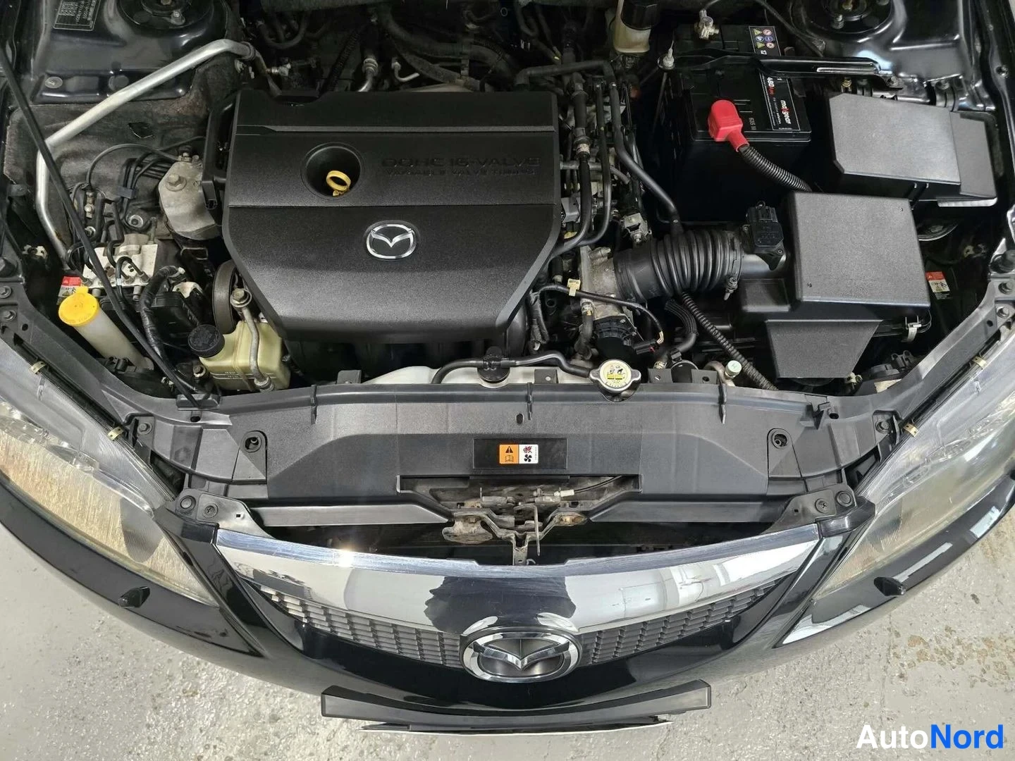 Muu: Mazdan moottoritila: DOHC 16V -moottorin suojakansi Mazda-emblemillä, akku ja ilmaputki näkyvät; etusäleikkö ja Mazda-tunnus kuvan alareunassa.