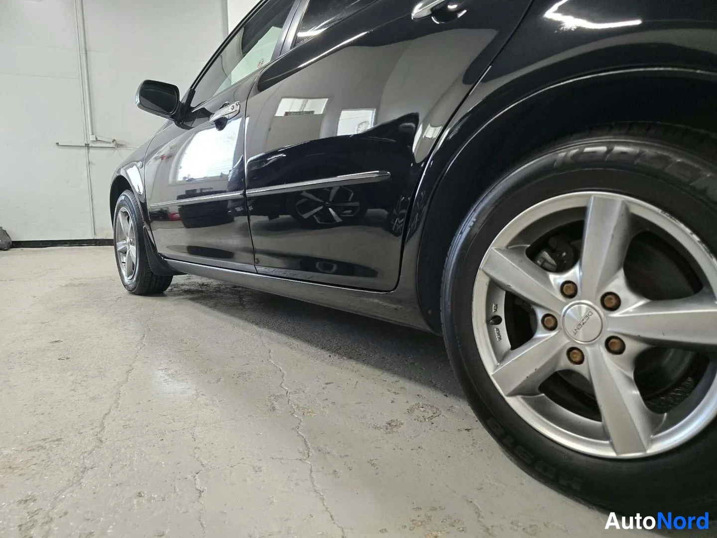 Sivulta: Mustan Mazda 6:n kylki ja takapyörä hopeisella viisipuolikkaalla vanteella kuvattuna lähikulmasta autotallin betonilattialla.