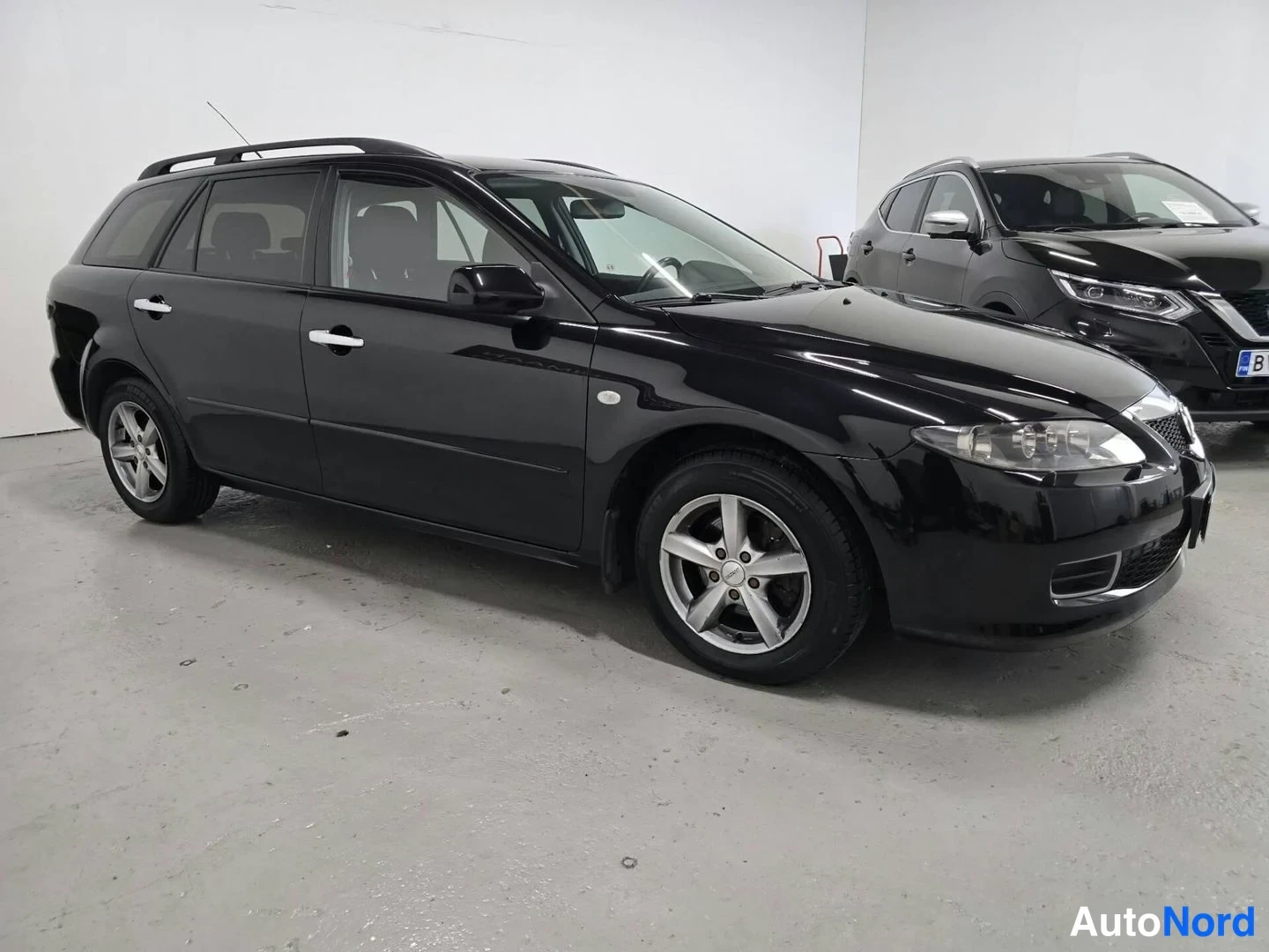 Etuviisto, nokka oikealle: Musta Mazda 6 -farmariauto kolmella neljäsosasta etukuvaan sisätilassa, etusäleikkö ja ajovalot näkyvät kuvan oikealla puolella.