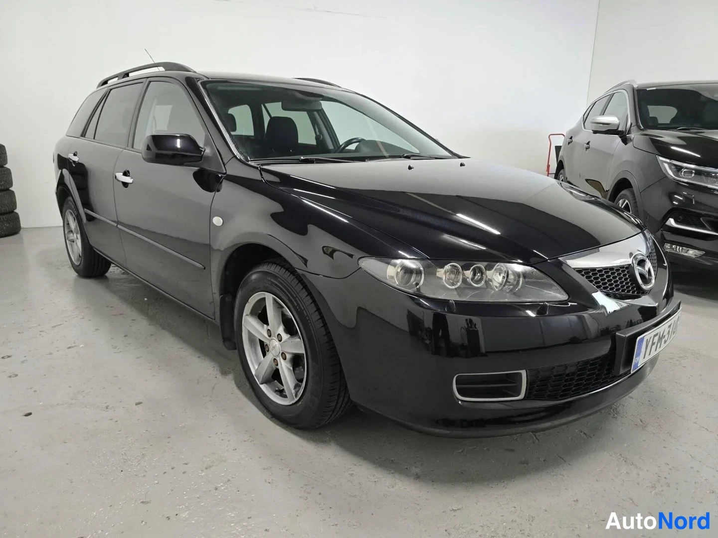 Etuviisto, nokka oikealle: Musta Mazda 6 -farmariauto etukulmasta sisätilassa, rekisterikilpi ja etusäleikkö näkyvät kuvan oikeassa laidassa.