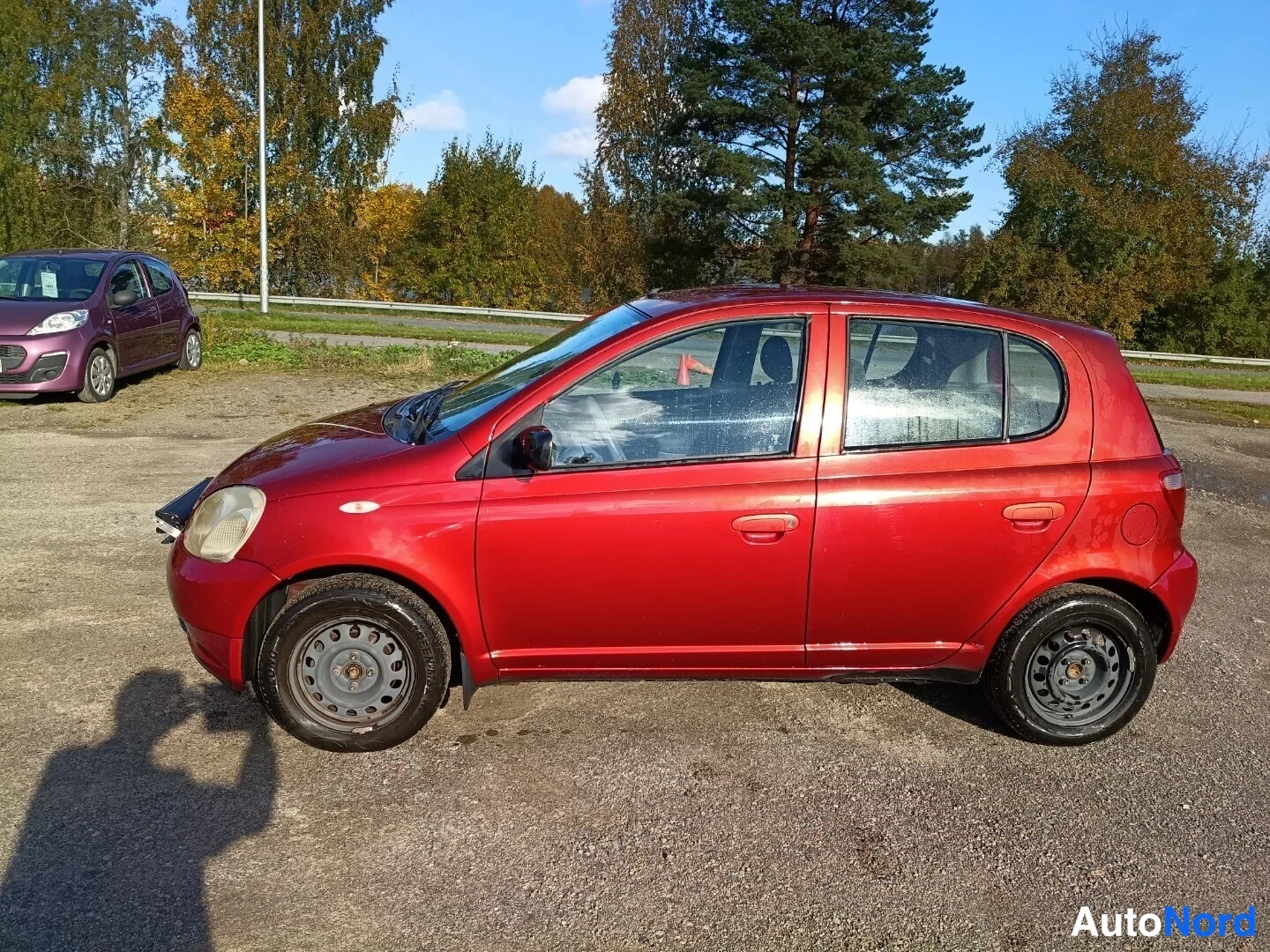 Etuviisto, nokka vasemmalle: Punainen Toyota Yaris sivulta vasemmalta, hiekkapintaisella parkkipaikalla