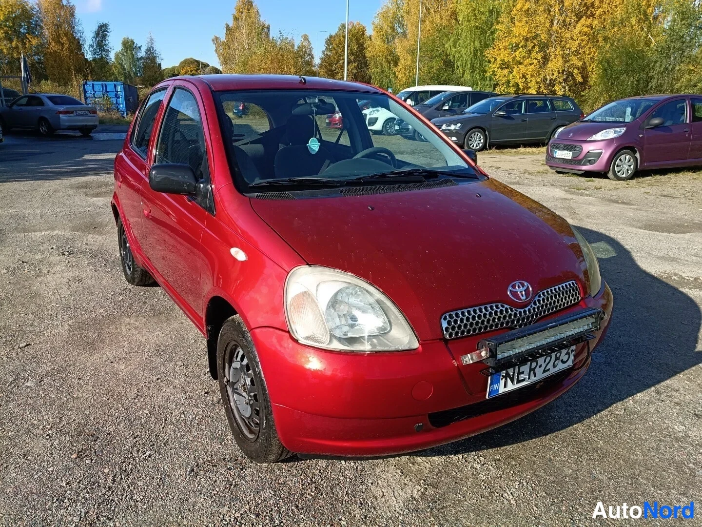 Etuviisto, nokka oikealle: Punainen Toyota Yaris etukulmasta oikealta, konepelti ja ajovalot näkyvissä, rekisteritunnus