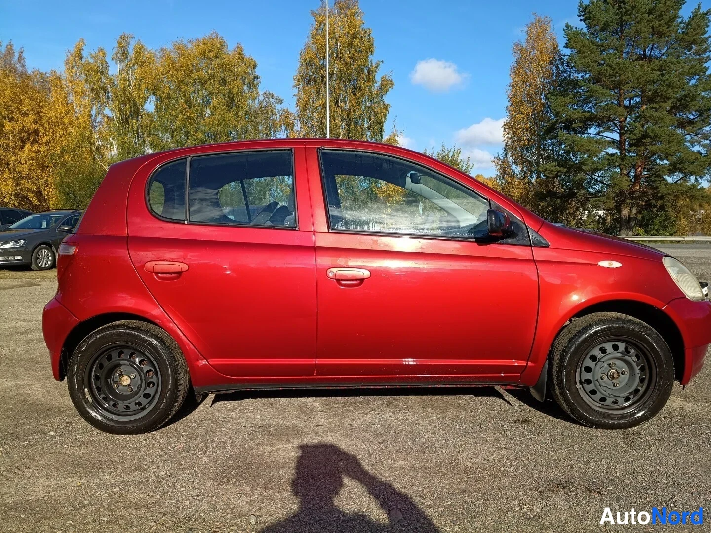 Sivulta: Punainen Toyota Yaris -henkilöauto sivulta kuvattuna, viistosti oikeanpuoleista keulaa kohti kallistunut profiili sorapysäköintialueella syksyisessä maisemassa.