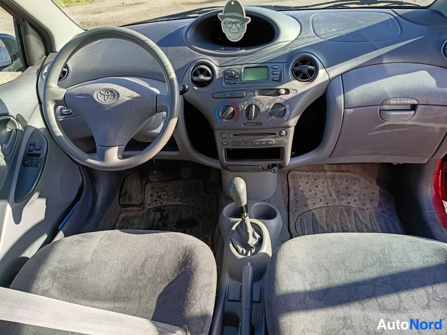 Edestä: Toyota Yarisin etuohjaamo ja kojelauta kuvattuna ovista: Toyota-merkkinen ohjauspyörä, analogiset mittarit, äänentoistolaitteet, ilmastointilaitteiden säätimet