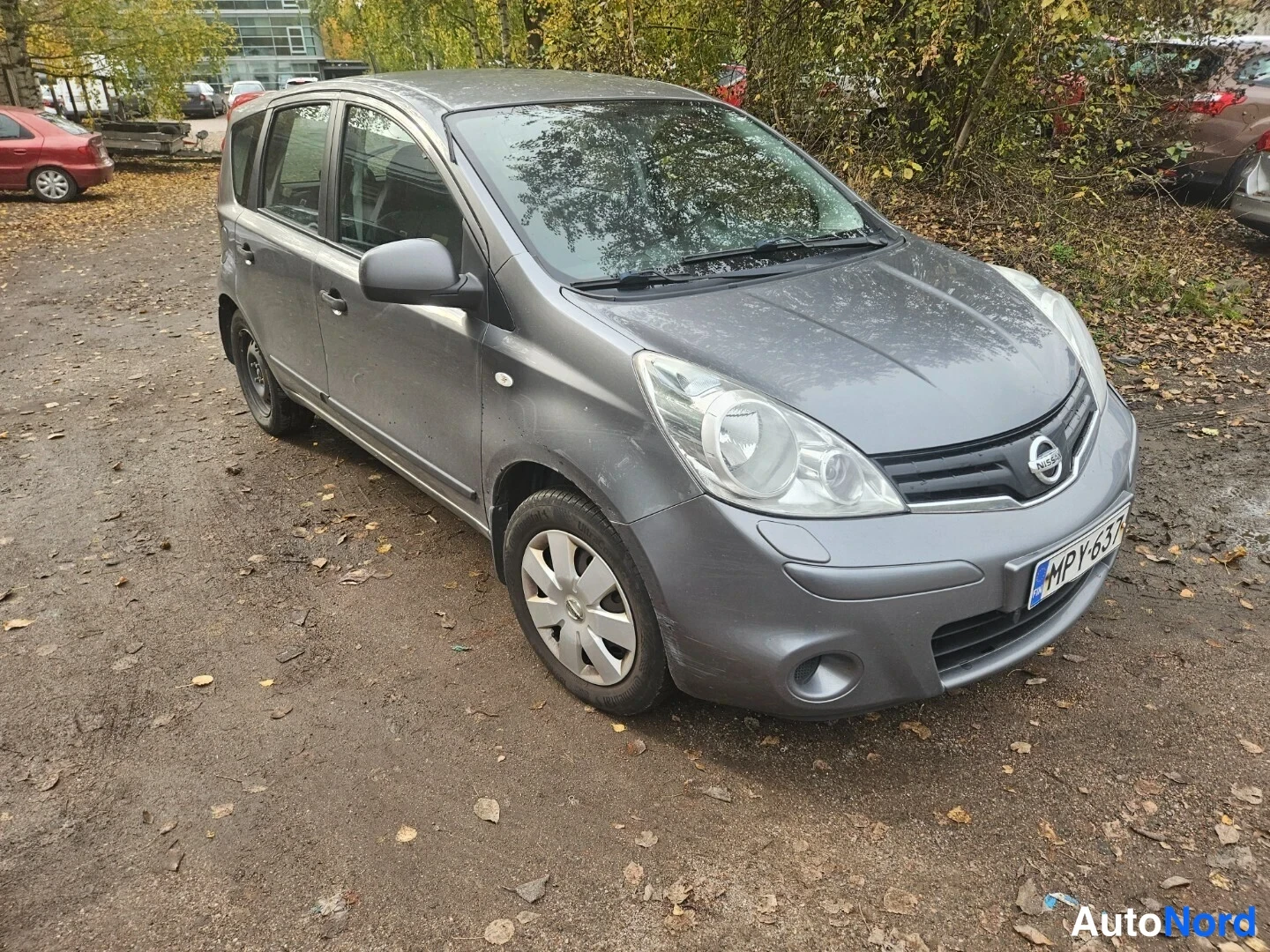 Etuviisto, nokka oikealle: Harmaa Nissan Note etukulmasta oikealta, rekisteritunnus, syyslehtien peittämällä parkkipaikalla.