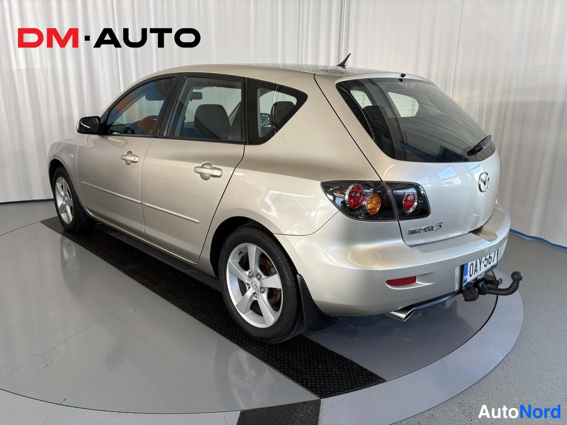 Takaviisto: Mazda 3:n takakulmanäkymä vasemmalta takaa; hopeavärinen koripaneeli, takavalaistus, rekisteritunnus, takakoukku kiinnitettynä.