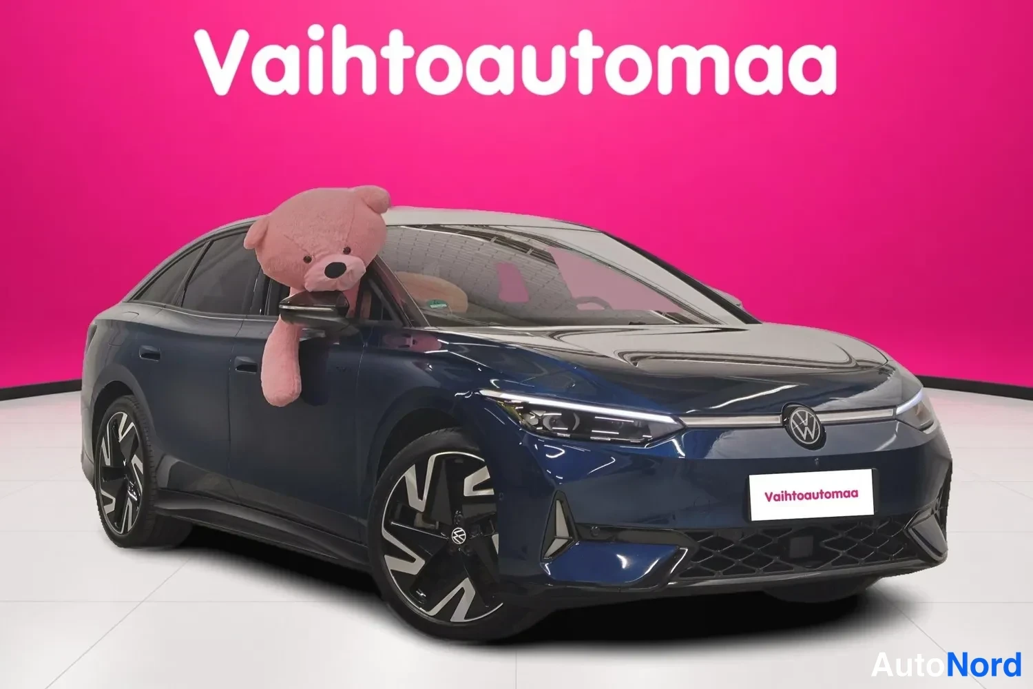 Etuviisto, nokka oikealle: Tummansininen Volkswagen ID.7 vinosti edestä oikealta kuvattuna; keula, kylki ja CCD-tyyppiset alumiinivanteet näkyvät, pinkissä studioympäristössä.
