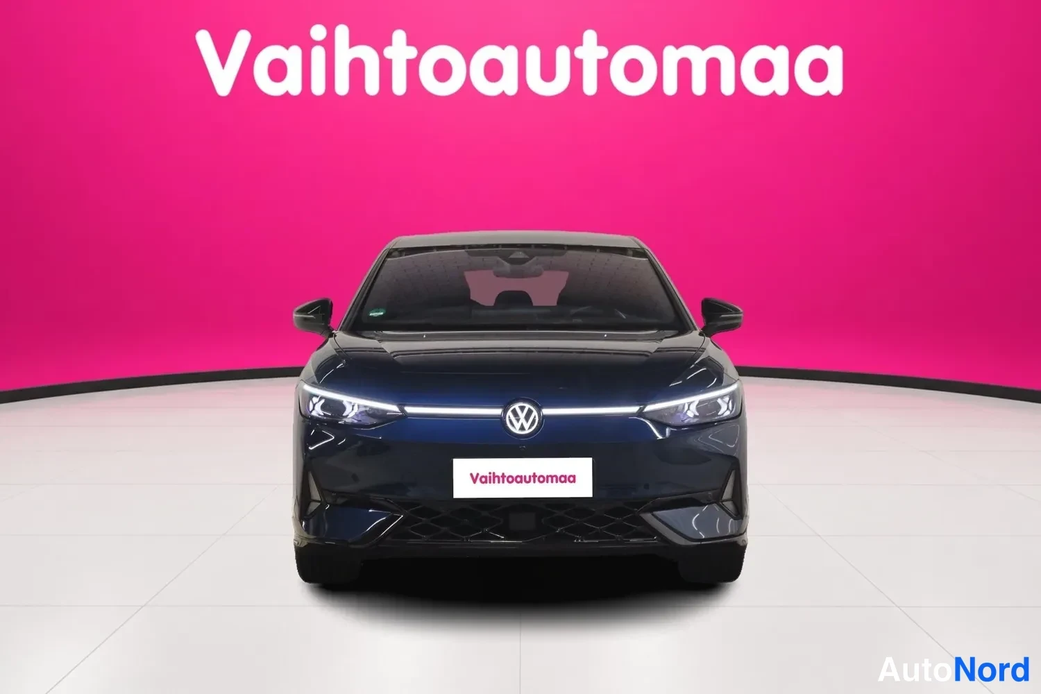 Edestä: Tummansininen Volkswagen ID.7 suoraan edestä kuvattuna; LED-päiväajovalot ja korinkestävä värivalotunnus näkyvät, pinkissä studioympäristössä.