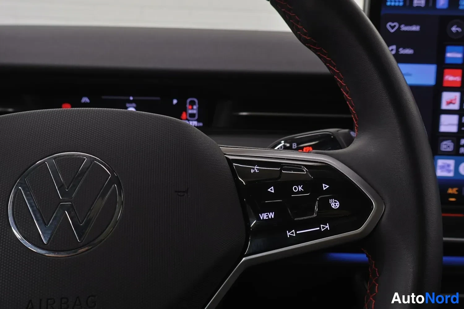 Yksityiskohta: Volkswagen-ohjauspyörän vasen puhelin, jossa infotainment-napit OK, VIEW ja äänikomennot, taustalla osittain näkyvä mittaristo ja punatiiminen ohjauspyörän reun