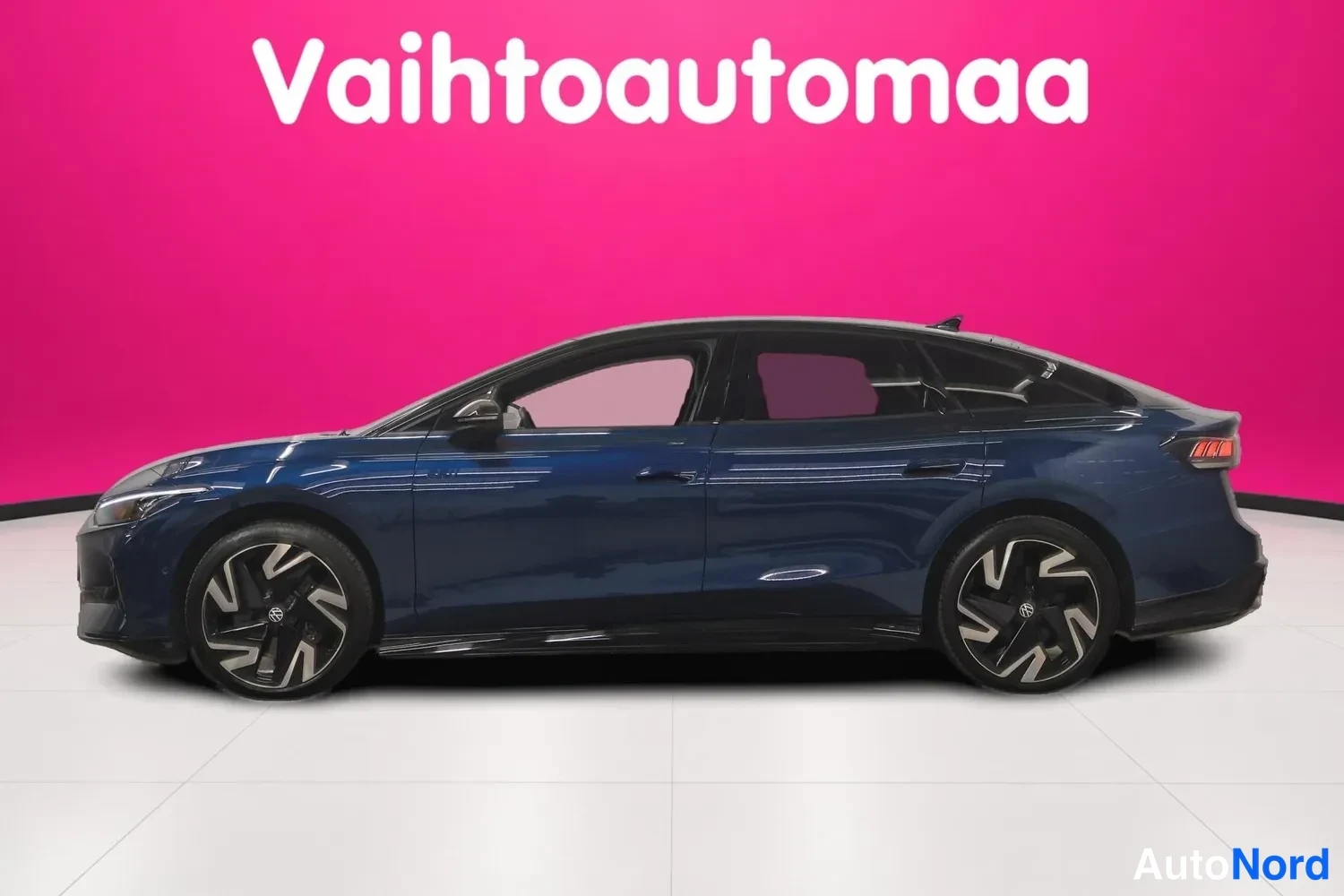 Sivulta: Tummansininen Volkswagen ID.7 sivulta kuvattuna; neliosaiset ovet, moniosaiset alumiinivanteet ja peräosien ääriviivat näkyvät, pinkissä studioympäristössä.