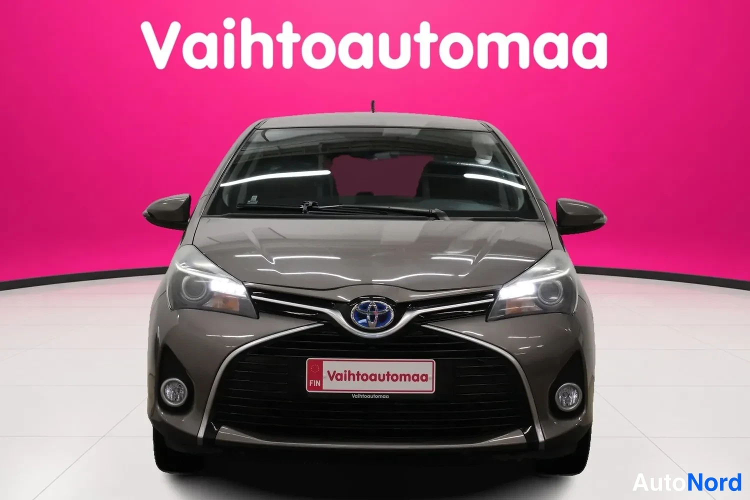 Edestä: Toyotan suora etunäkymä: harmaanruskea yaris, tummat ajovalot ja pinkin taustan edessä.