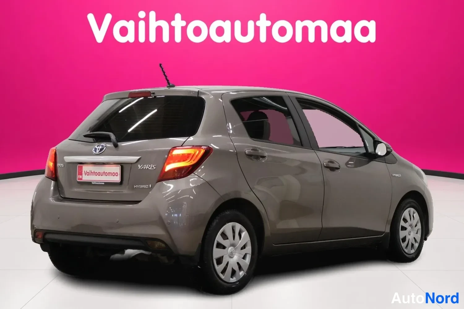 Takaviisto: Toyota Yaris -auton takakulma vasemmalta, tummanharmaa koristus, Hybrid-tunnus takaluukussa, Vahtoautomanalo liila-taustaa vasten.