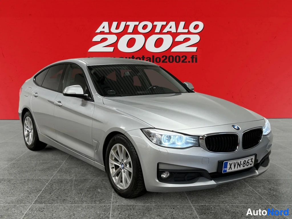 Etuviisto, nokka oikealle: Hopeanvärinen BMW 3 Gran Turismo -viistoperäauto edestä oikealta kulmasta kuvattuna, rekisterikilpi, punainen tausta.