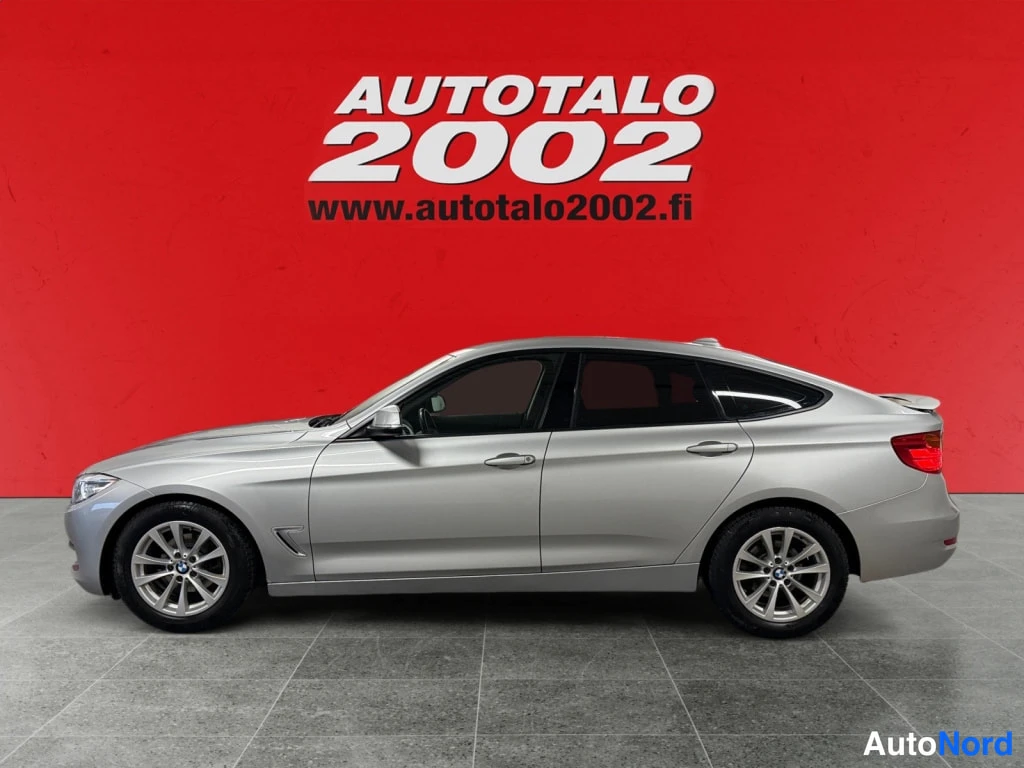 Etuviisto, nokka vasemmalle: Autotalo 2002 -kyltillä BMW 318 Gran Turismo, sivulta oikealta kuvattu; etummainen ajovalo näkyy kuvan vasemmassa reunassa ovien ohella. Auton kori on