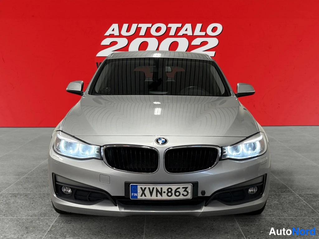 Edestä: Autotalo 2002 -kyltillä BMW 318 Gran Turismo suoraan edestäpäin; ajovalot ovat päällä, etusäleikössä BMW-merkki ja rekisterikilpi lukee. Kori on hopeanvärinen.