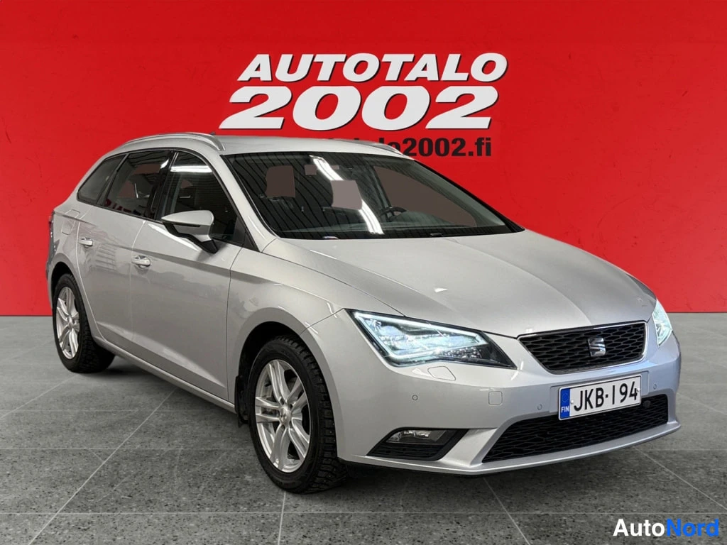 Etuviisto, nokka oikealle: Hopeanvärinen Seat Leon ST -farmariauto esillä punaisella taustalla, kuvattu etuviistöstä; rekisterikilpi.
