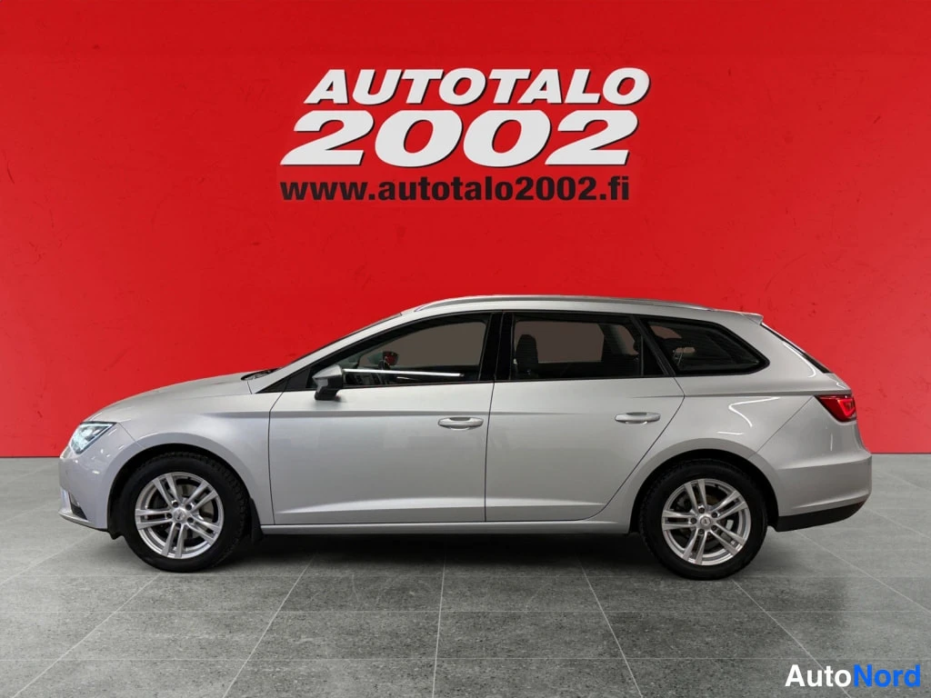 Takaviisto: Autotalo 2002 -mainos taustalla; harmaa Seat Leon ST -farmariauto sivulta kuvattuna, kattonivelten (kattokiskojen) päälli ja hopeiset kevytmetallivanteet näkyvä