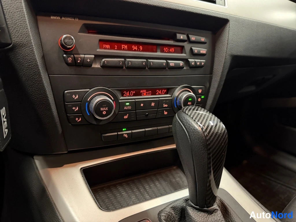 Muu: Kojelaudan keskikonsolin lähikuva: BMW Business CD -radio näyttää FM 94.9 -kanavaa, alla automaattinen kaksivyöhykeilmastointi (24°) ja varaustaso. Hiilikuituin