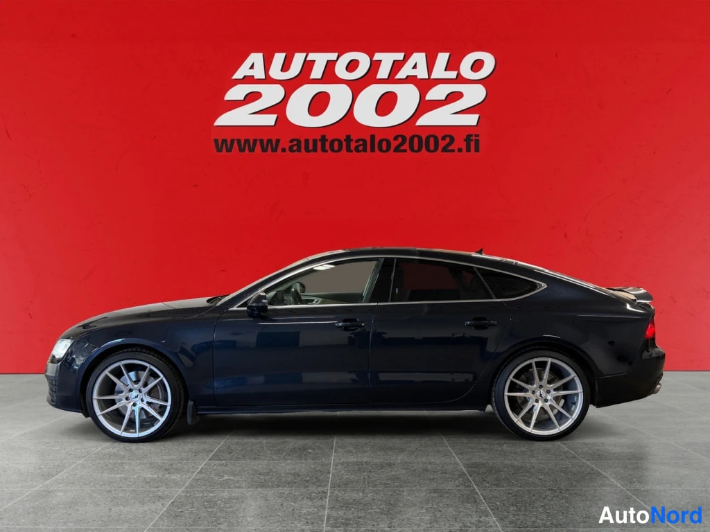Sivulta: Sinimusta Audi A7 sivusuunnassa punaisella taustalla, hopeiset kevytmetallivanteet.