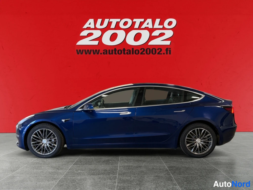 Sivulta: Sivukuvassa tummansininen Tesla Model 3 -henkilöauto kuvattuna sivulta punaisella taustalla.