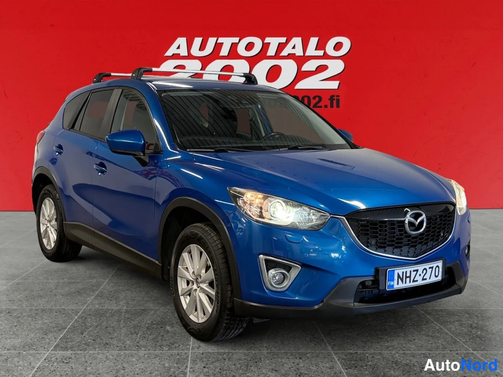 Etuviisto, nokka oikealle: Sininen Mazda CX-5 etuviistosta oikealta, kattokaiteet ja rekisterikilpi näkyvissä.