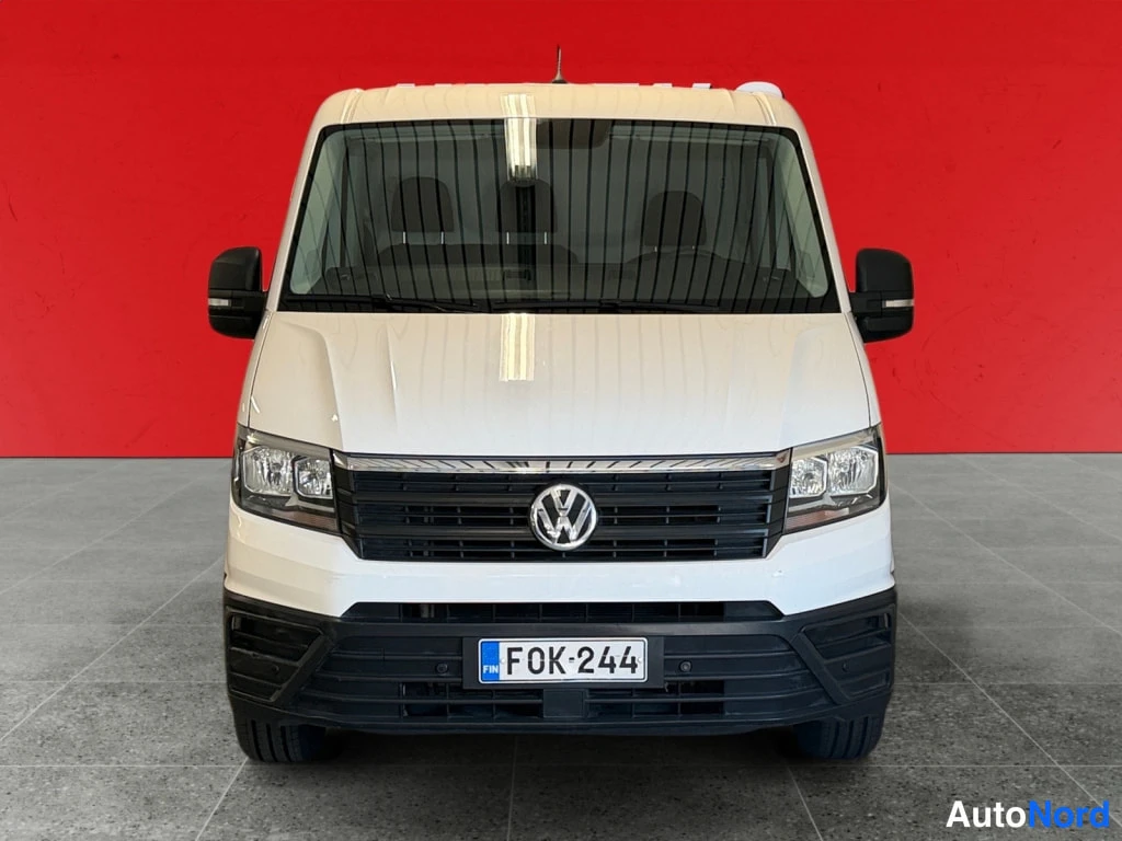 Edestä: Valkoisen pakettiauton etunäkymä, VW-merkki ja rekisteritunnus, punainen tausta.