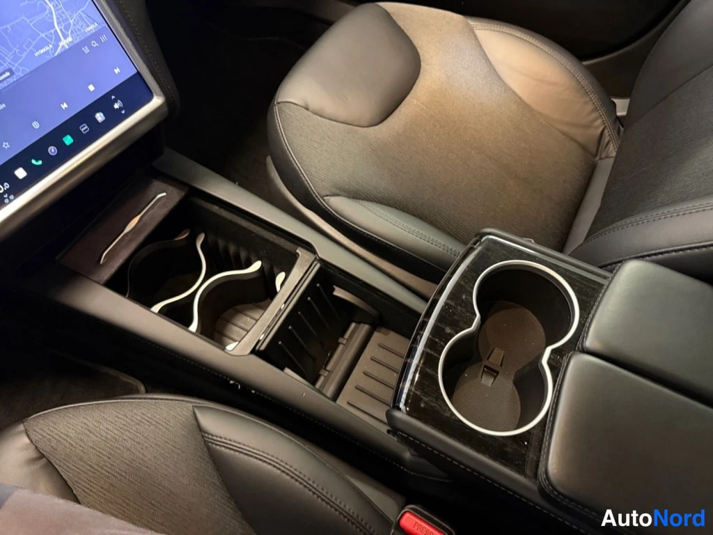Edestä: Sisäkuva Tesla Model S -auton etuosasta: musta/p harmaa nahkaverhoilu, kupeet, musta keskitaso cupholderineen ja taitettu hylly.