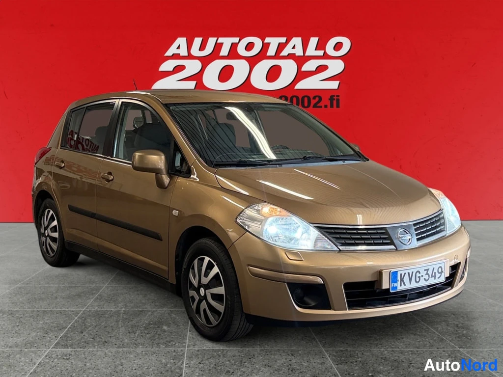 Etuviisto, nokka oikealle: Kultaisenruskean Nissan Tiida -viistoperän etukolmannelta sivulta oikeaan kulmaan kuvattuna, punainen Autotalo 2002 -tausta.