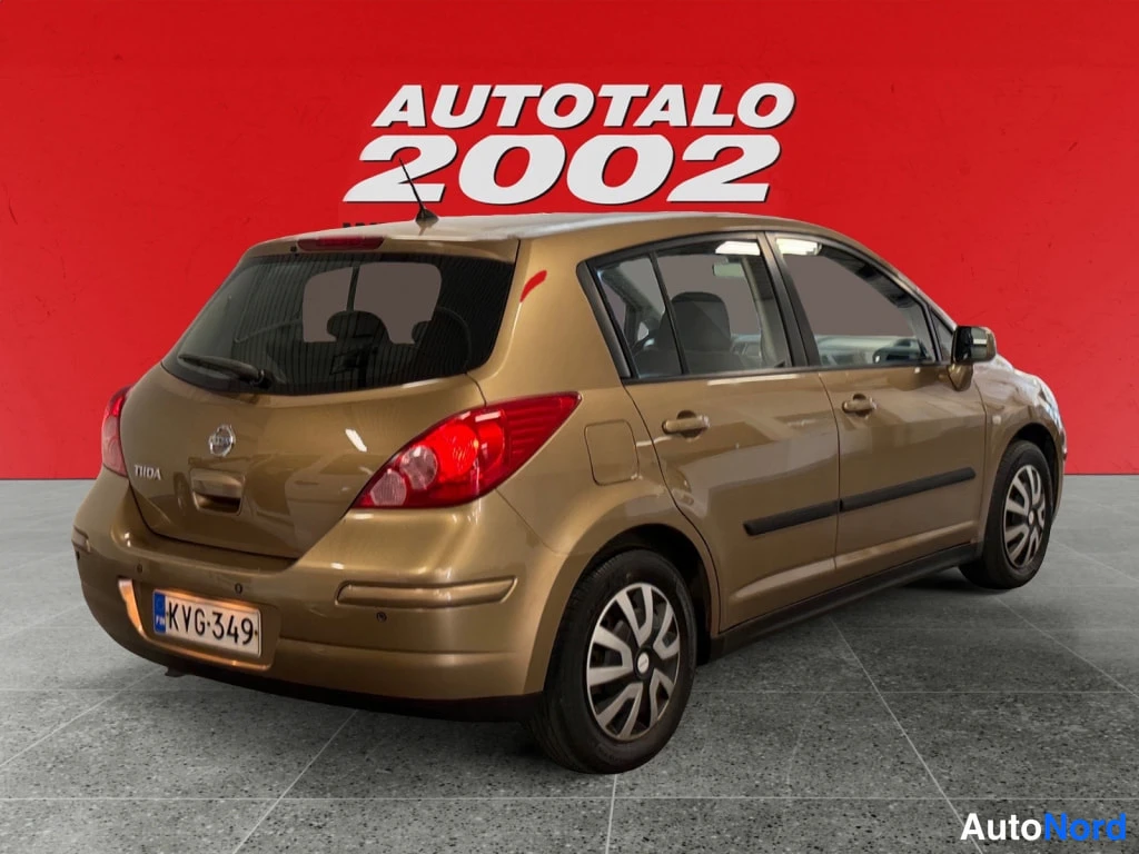 Takaviisto: Kultaisenruskean Nissan Tiida -viistoperän takakolmannelta vasemmalle kuvattuna, punainen Autotalo 2002 -tausta.