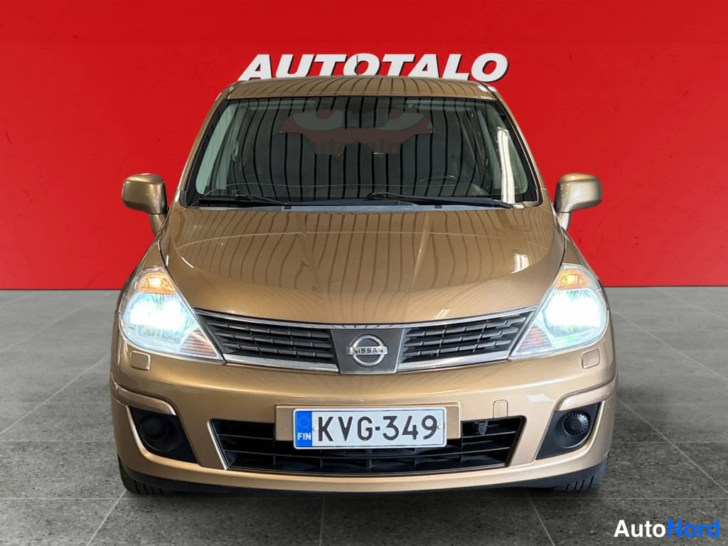Edestä: Kultaisen Nissan Tiida -auton keulakuva suoraan edestä, etuvalat päällä, rekisterikilpi, punainen tausta.