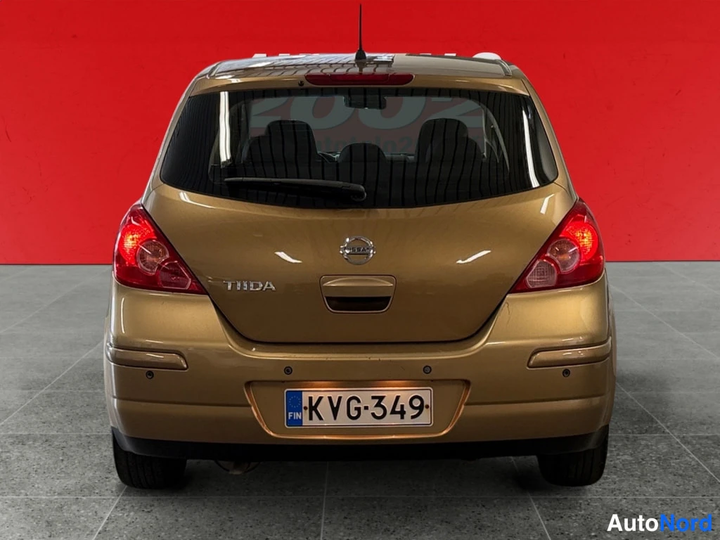 Takaa: Kultaisen Nissan Tiida -auton perä suoraan takaa, TIIDA-merkintä, takavalot, rekisterikilpi, punainen tausta.