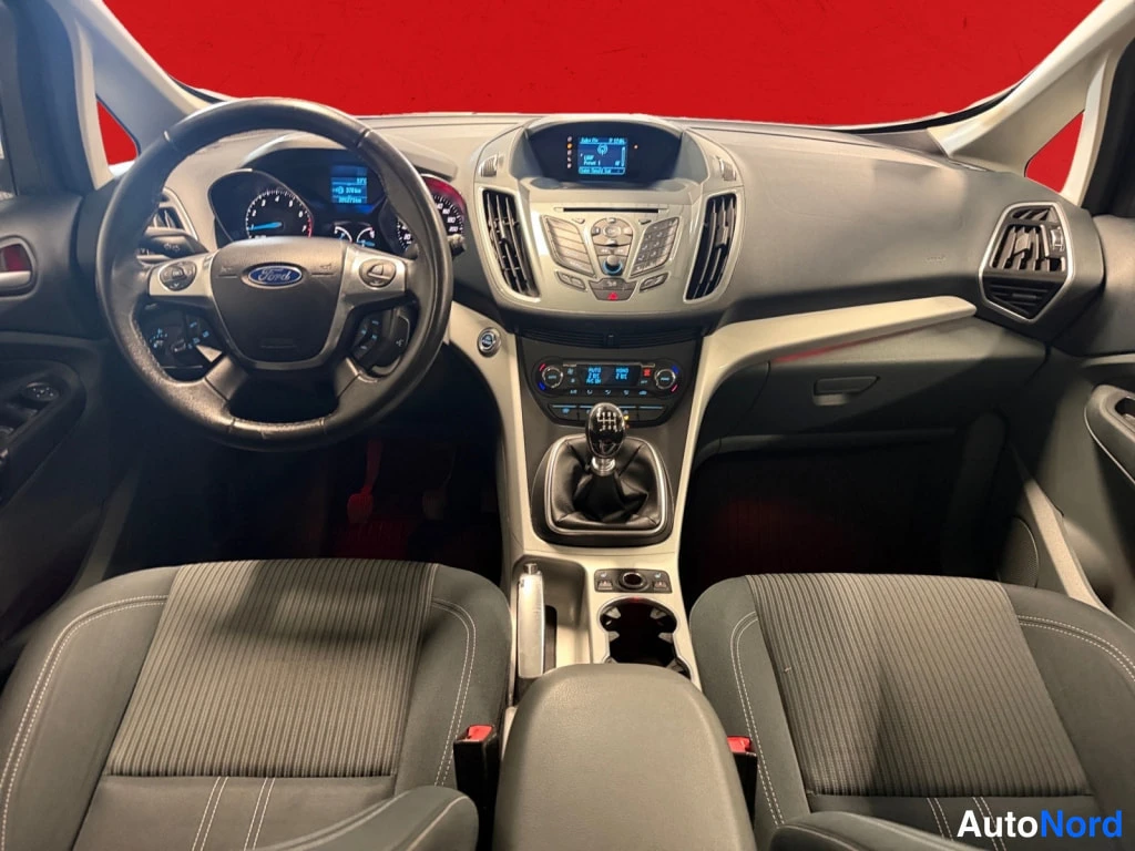 Edestä: Auton kojelauta edestäpäin: Ford-ohjauspyörä, analogiset mittarit, keskikonsolin näyttö ja radiopaneeli, manuaalivaihtaja, kaksi etuistuinta tummassa kankaassa.