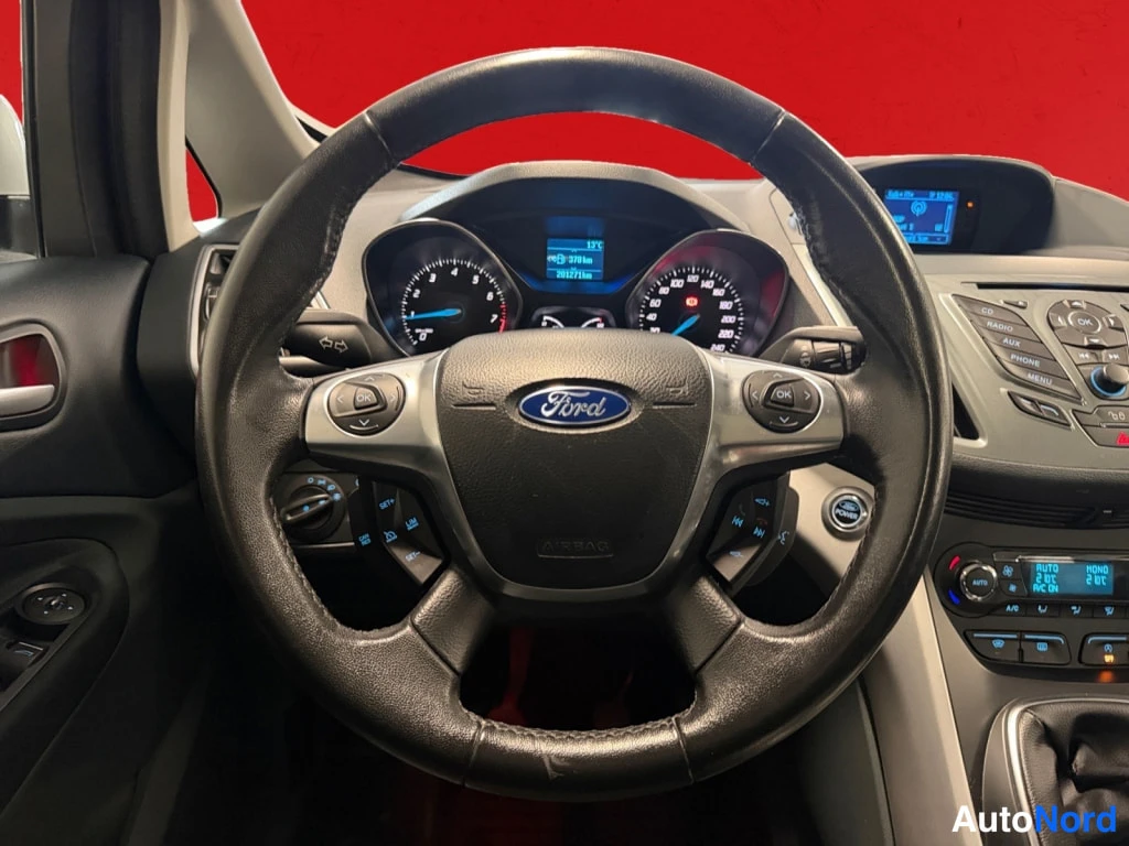 Muu: Ohjaamo näkyvä kuvattuna kuljettajan paikalta: Ford-merkkinen ohjauspyörä, mittaristo ja kojelaudan näytöt punaisella taustalla.