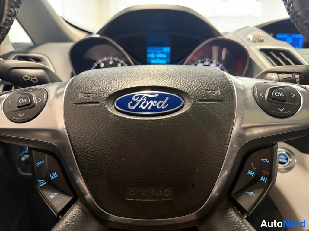 Yksityiskohta: Ford-ohjauspyörän keskusta AIRBAG-tekstillä, sininen Ford-merkki sekä monitoiminapit (OK, SET+, LIM, SET-, puhelin- ja ääniohjausnapit); mittariston näyttö näky