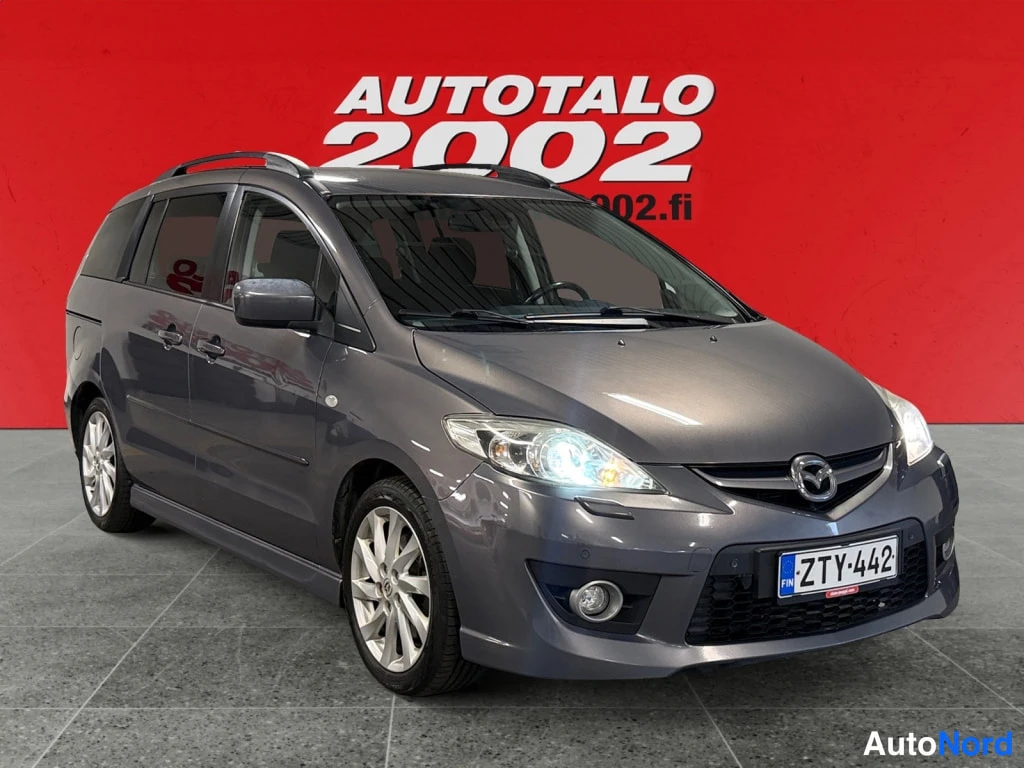 Etuviisto, nokka oikealle: Tummanharmaa Mazda 5 -tilausauto vinosti etuoikealta kuvattuna, etupään ja oikea kylki näkyvät, rekisteri.