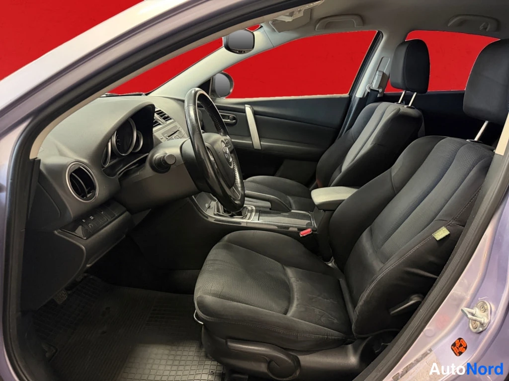 Yksityiskohta: Mazda 6:n etuistuintila sivulta avoimen kuljettajan oven kautta kuvattuna: mustat kankaisistuimet, korkea keskikonsoli ja Mazda-ohjauspyörä, taustalla punainen