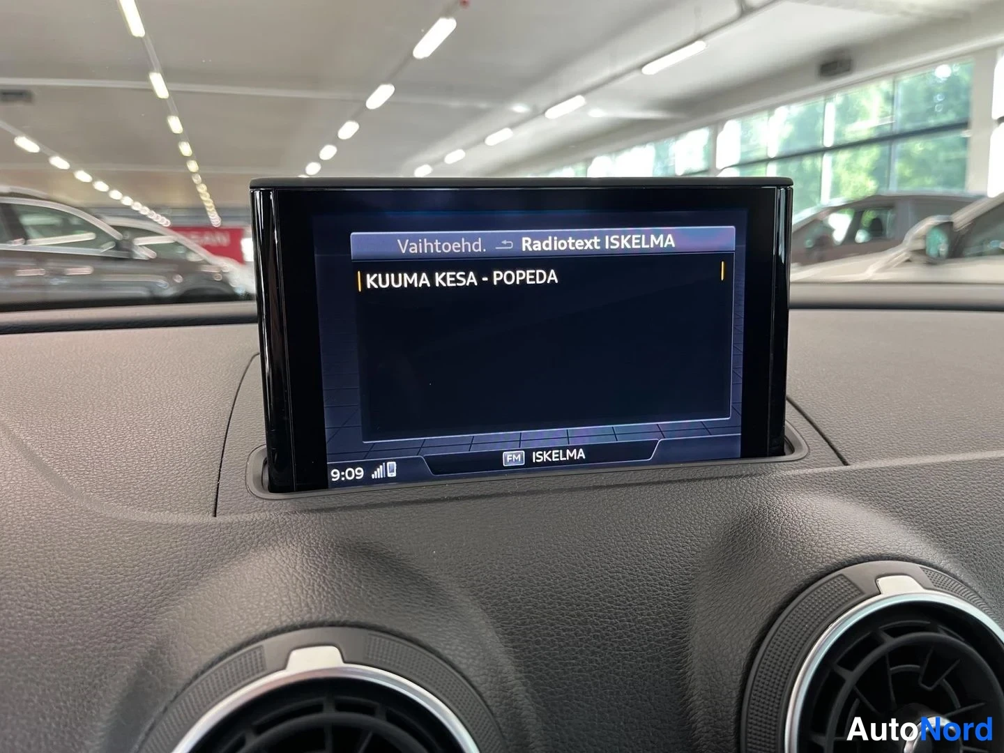 Muu: Sisätilakuva: keskikäyttöpaneelin näyttö näyttää ISKELMA-radioaseman radiotekstin 'KUUMA KESA - POPEDA', kellonaika 9:09, auto showroomissa.
