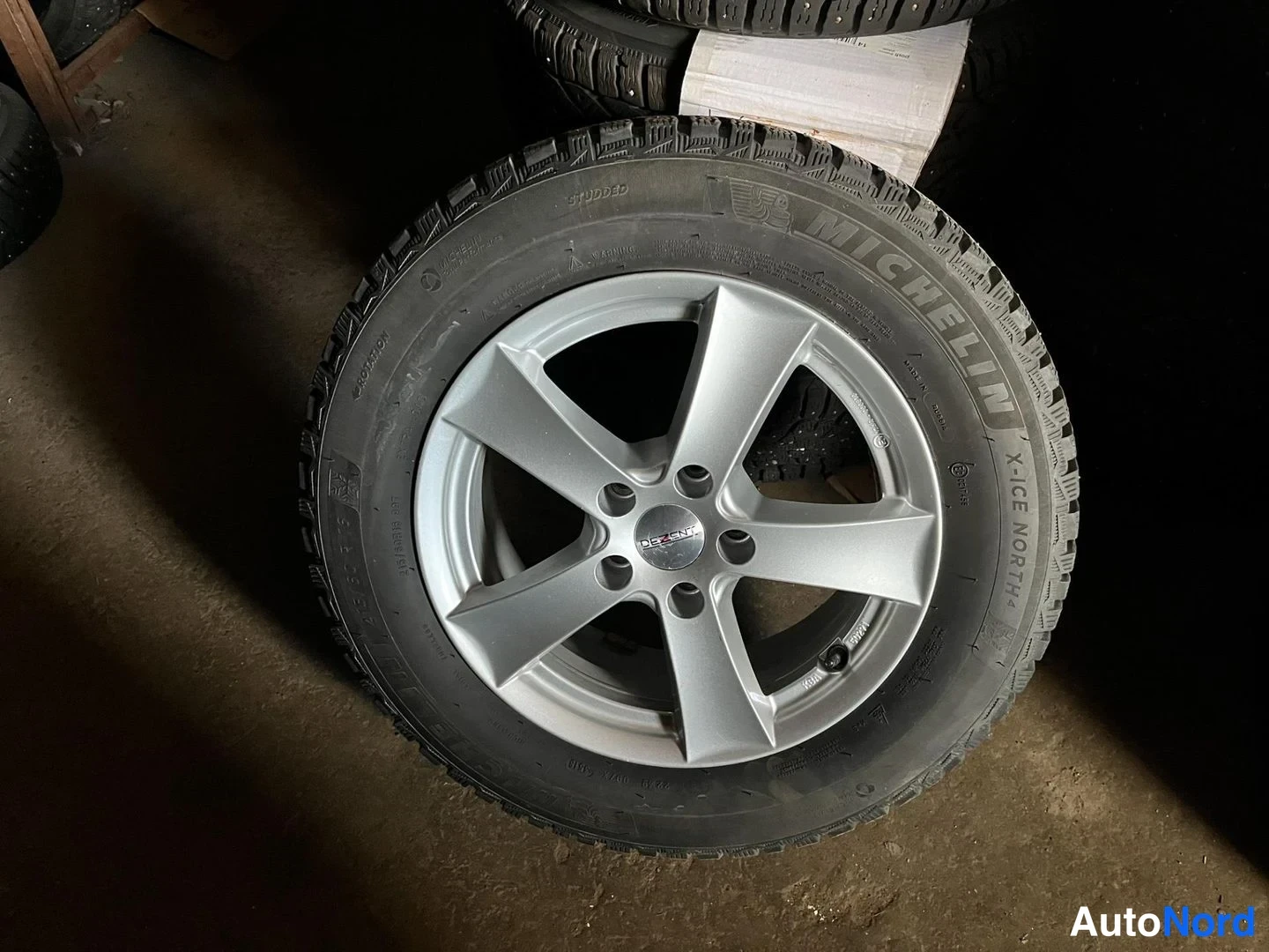 Yksityiskohta: Yksittäinen metallinvärinen viisipeittopyörä, jossa Michelin X-ICE North -nastarengas, seisoo varaston betonilattialla.