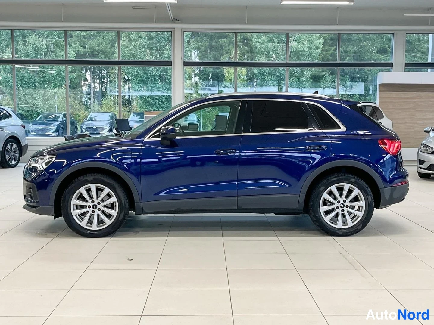 Etuviisto, nokka vasemmalle: Audi Q3 -maastoauto vasemmasta etukulmasta, tummansininen koripaneeli, hopeat vanteet ja tummat ikkunat showroomissa.