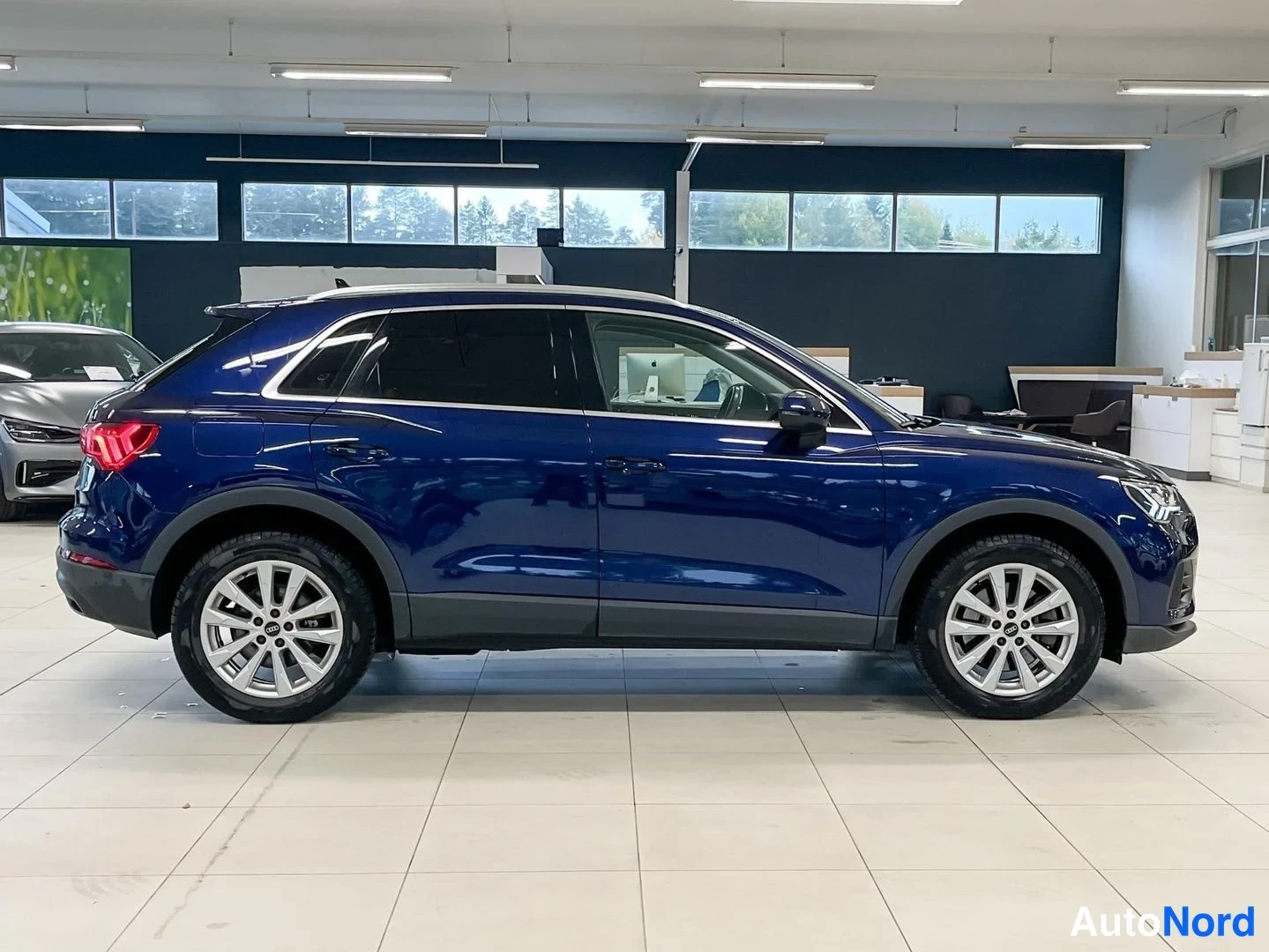 Takaviisto: Sinisenäinen Audi Q3 -maasturin taka-kolmasosan kuvakulma autonhallissa; sivuprofiili, takavalot ja hopeiset vanteet näkyvät.