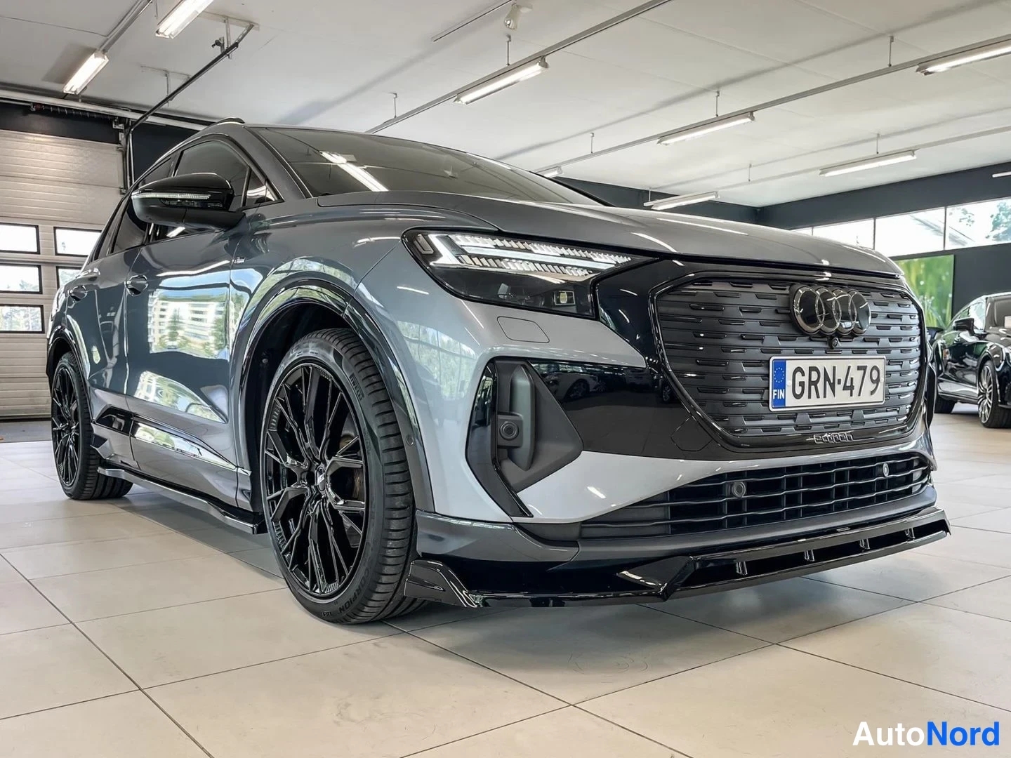 Etuviisto, nokka oikealle: Audi Q4 e-tron -maasturin etukulmanäkyvää sisätiloissa, harmaan korin ja mustien vanteiden näkyvä, rekisteritunnus.