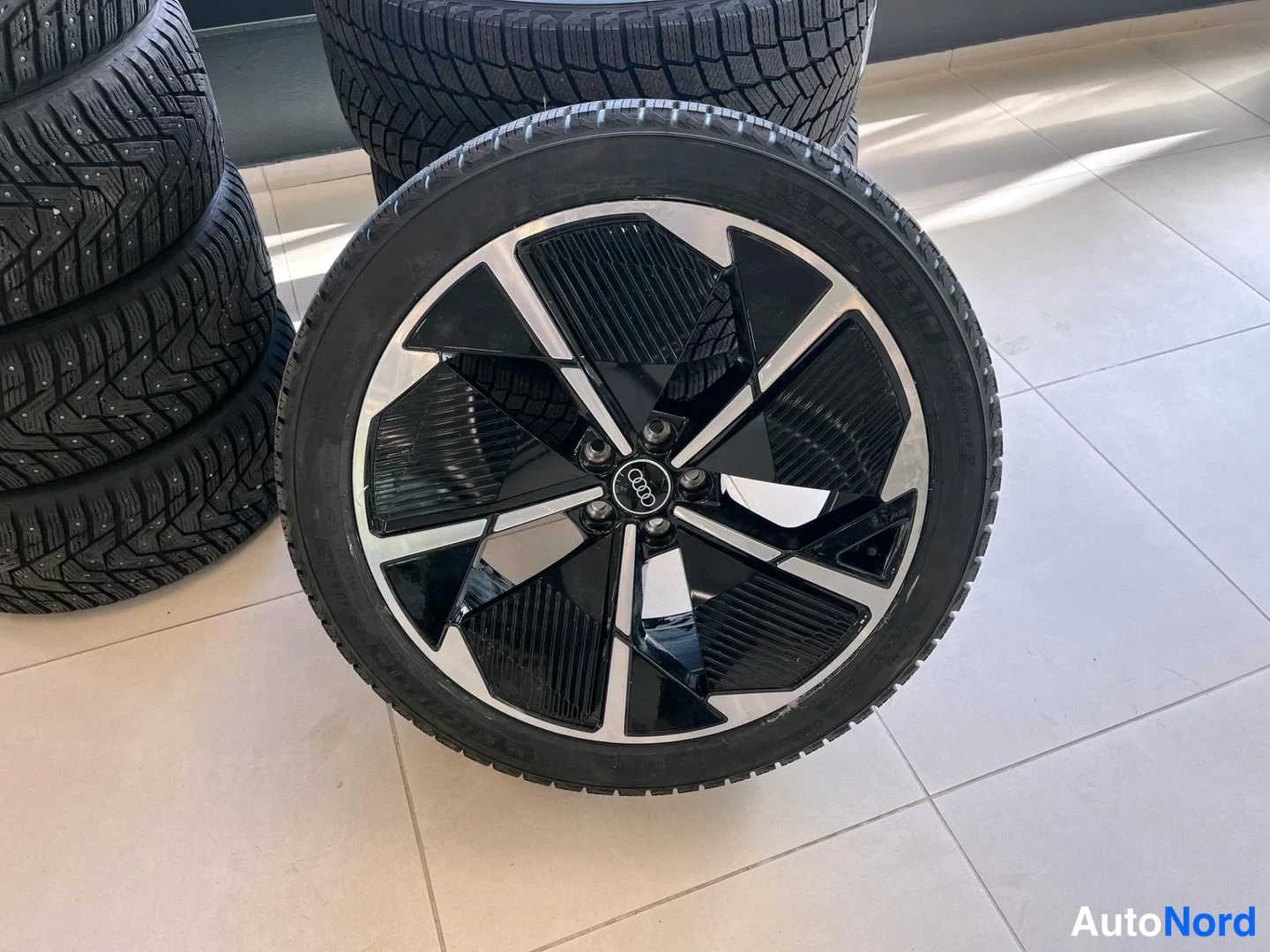 Yksityiskohta: Audi-merkitty talvirengas musta-hopeisella alumiinivanteella lattialla, taustalla muita rengassettejä.