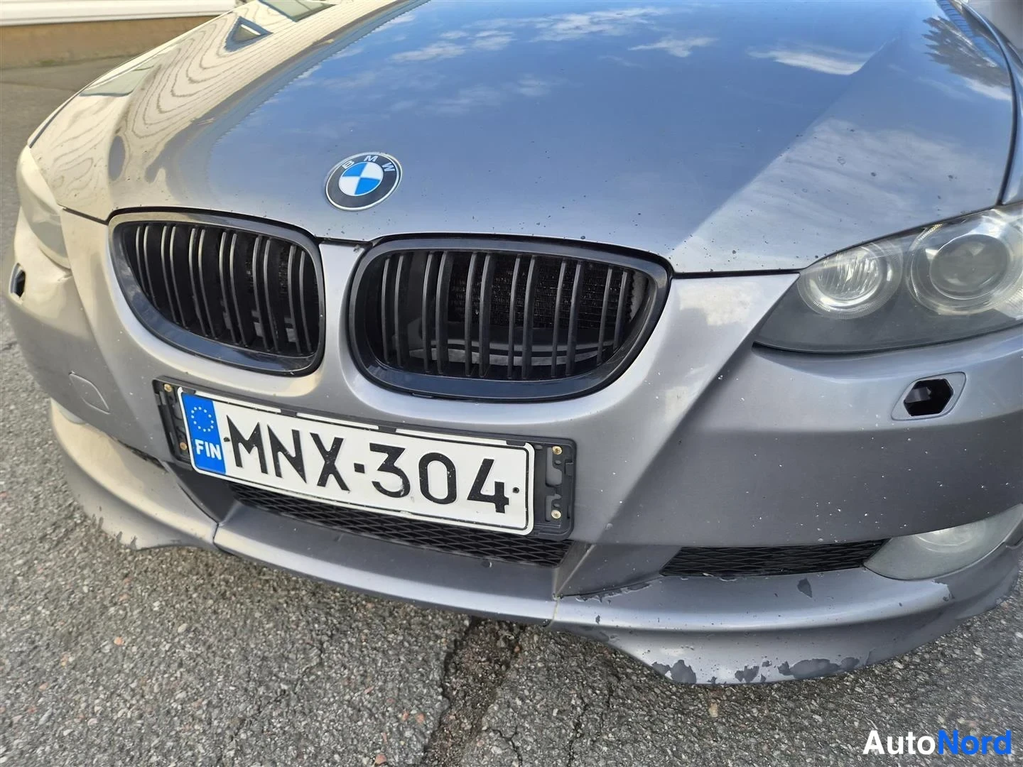 Edestä: BMW:n etuosa lähikuvassa: tummat munuaiset, etvalot ja rekisterikilpi, puskurin alareunassa lohkeilevaa pintaa.