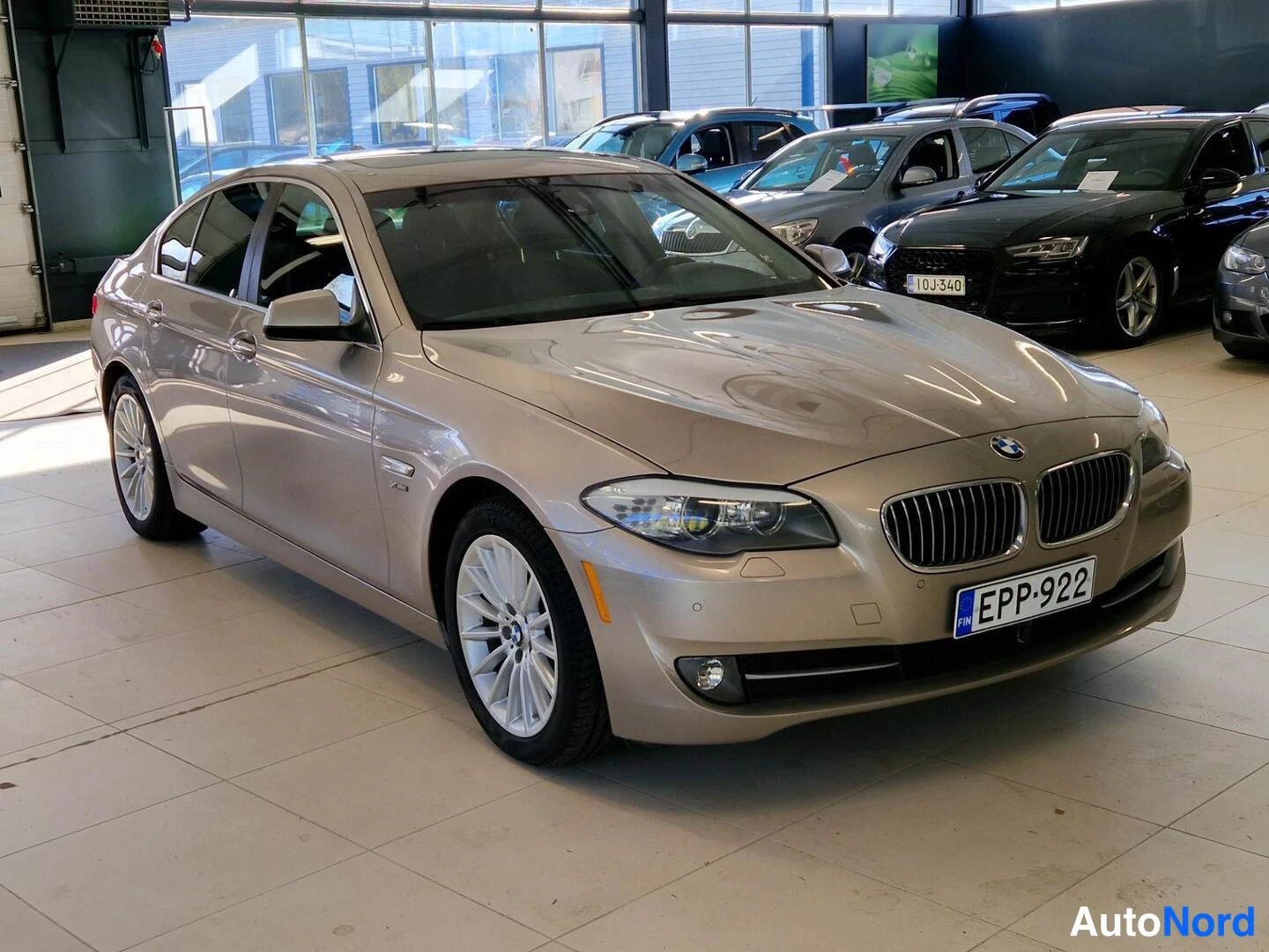 Etuviisto, nokka oikealle: Kultainen BMW 5-sarjan sedanin etukulmakuva sisätiloissa, keulalla rekisterikilpi.