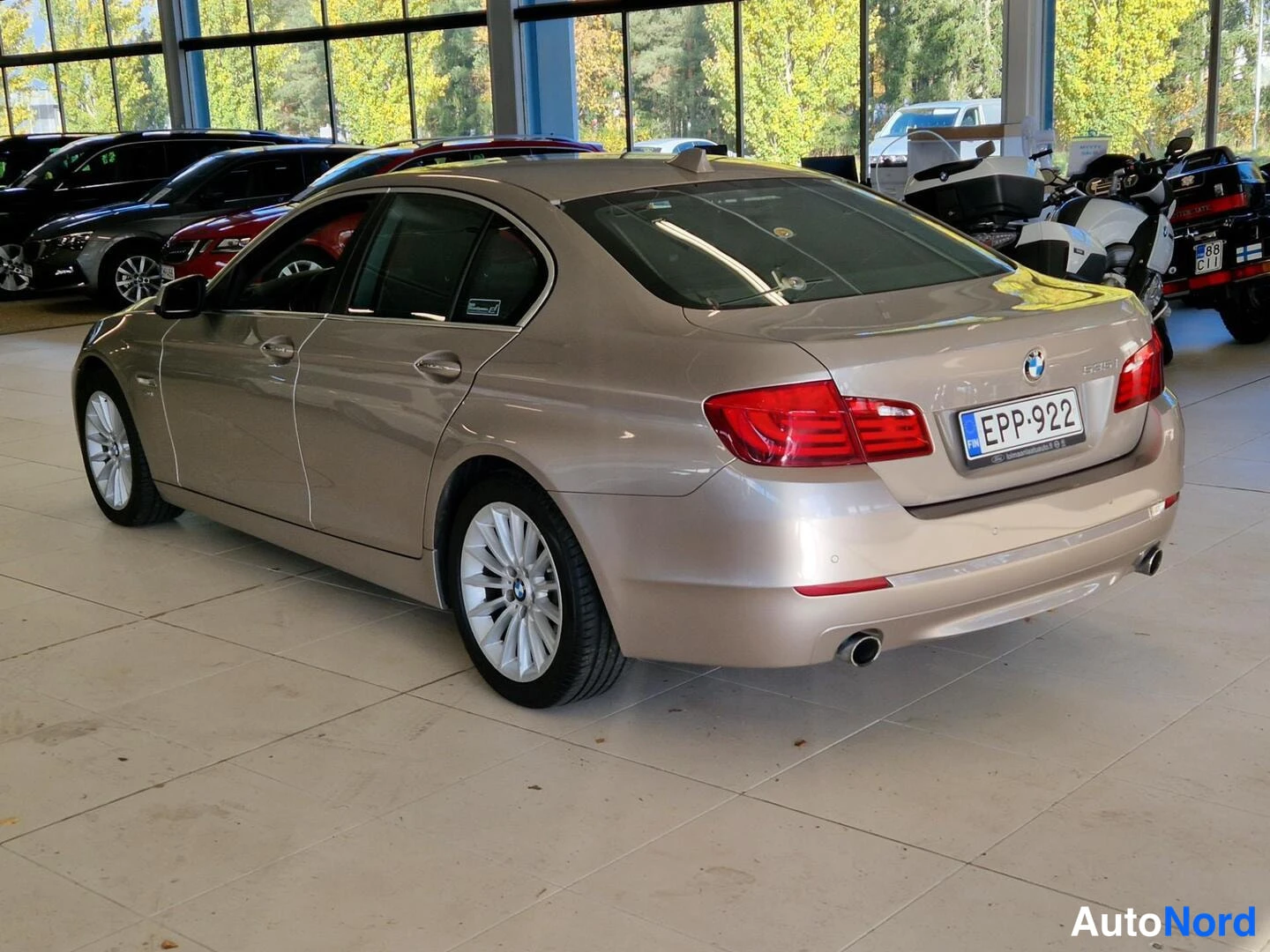 Takaviisto: Kultainen BMW 5-sarjan sedanin takakulmakuva sisätiloissa, takana rekisterikilpi.
