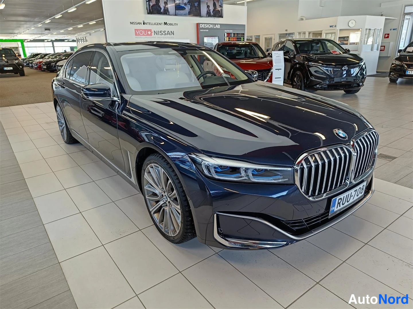 Etuviisto, nokka oikealle: Tummansininen BMW-sedan etukulmasta vasemmalle kallellaan, rekisterikilpi, esityshallissa.