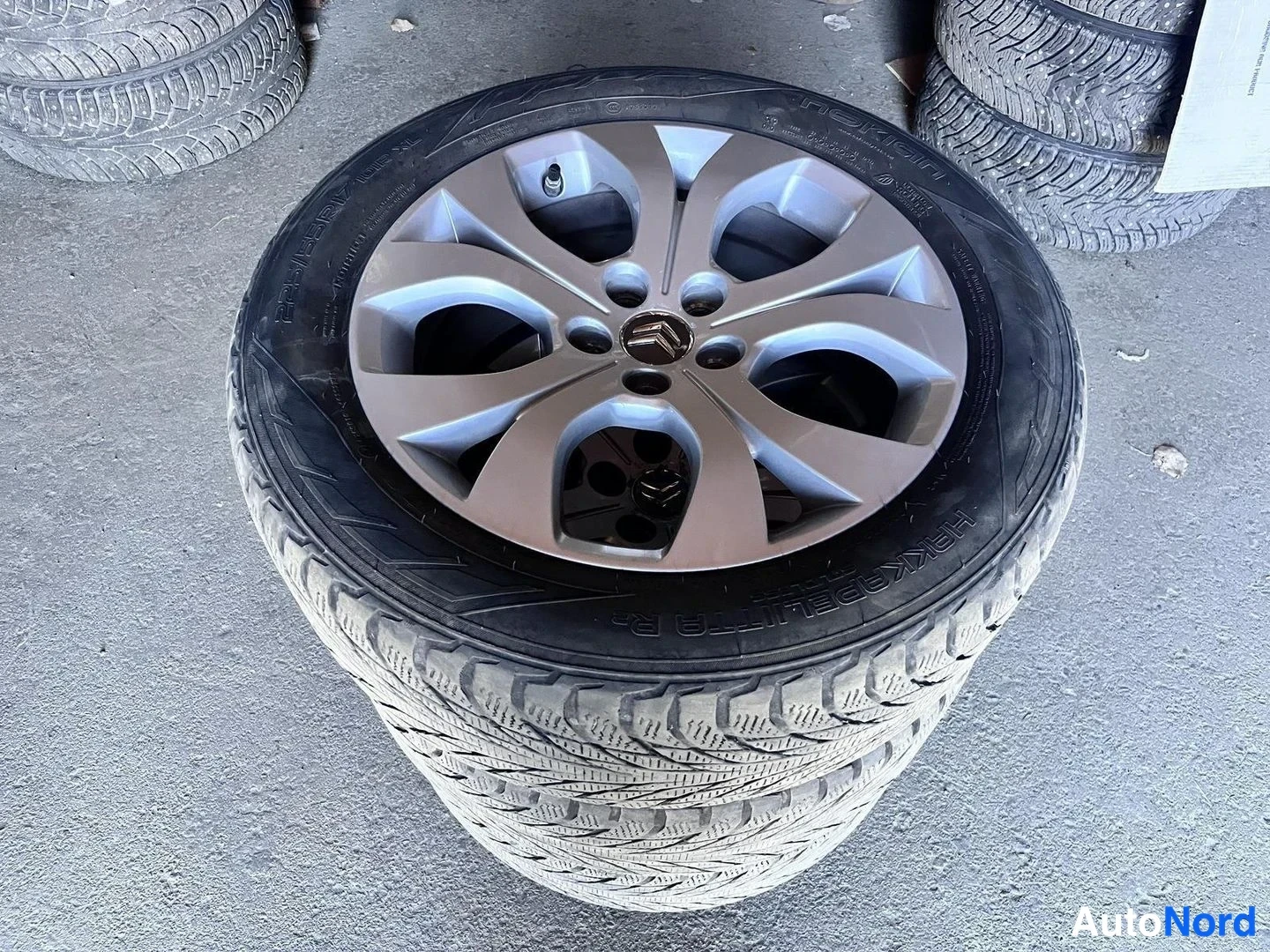 Yksityiskohta: Nokian Hakkapeliitta R2 -talvirengas vanteella, jossa Citroënin merkki, renkaita pinossa ja maassa.