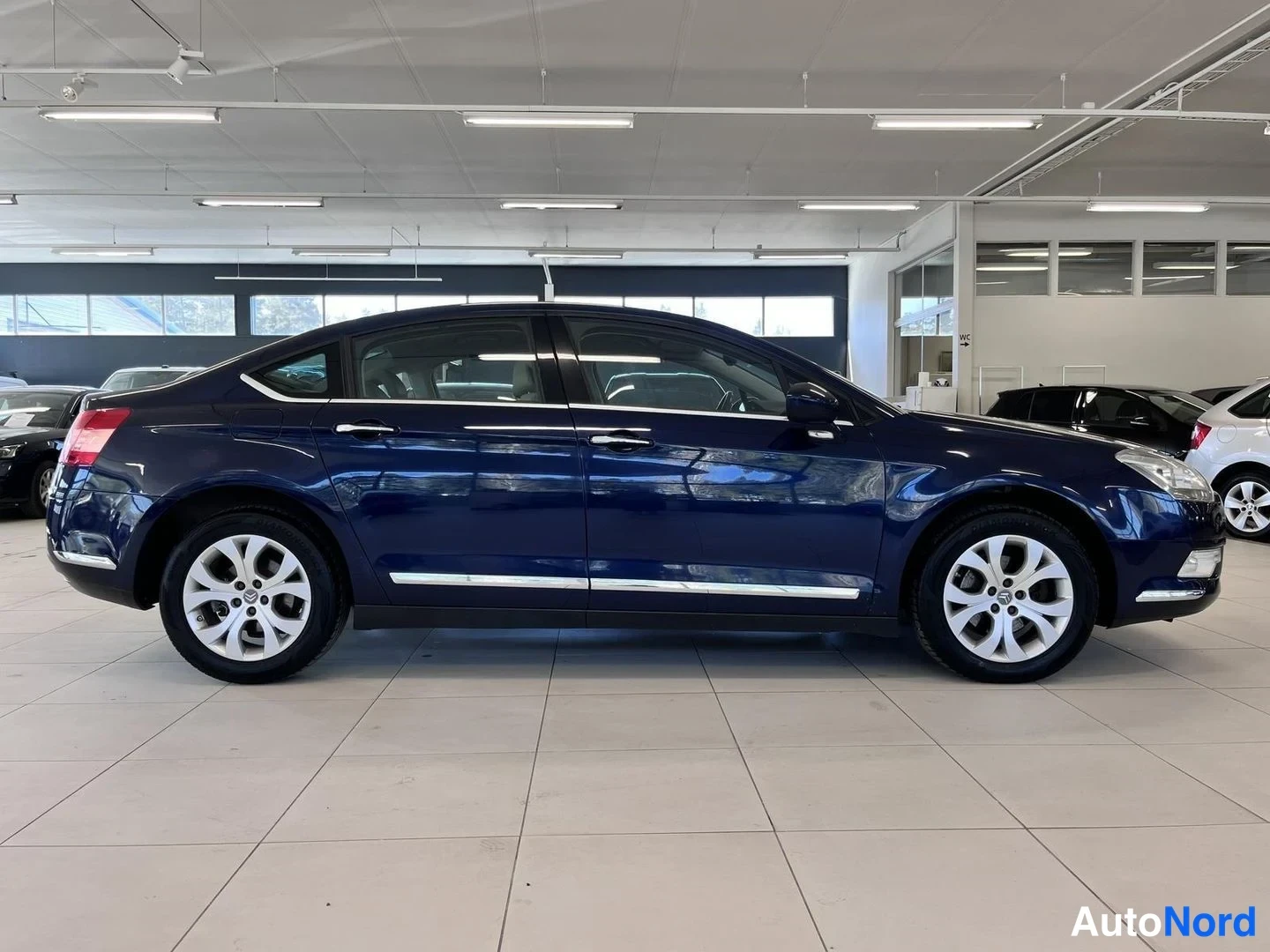 Sivulta: Tummansinisen Citroën C5:n vasen sivuprofiili autonosaliassa; neljä ovea, kromilistat ovien alareunassa ja hopeanväriset peltivanteet.
