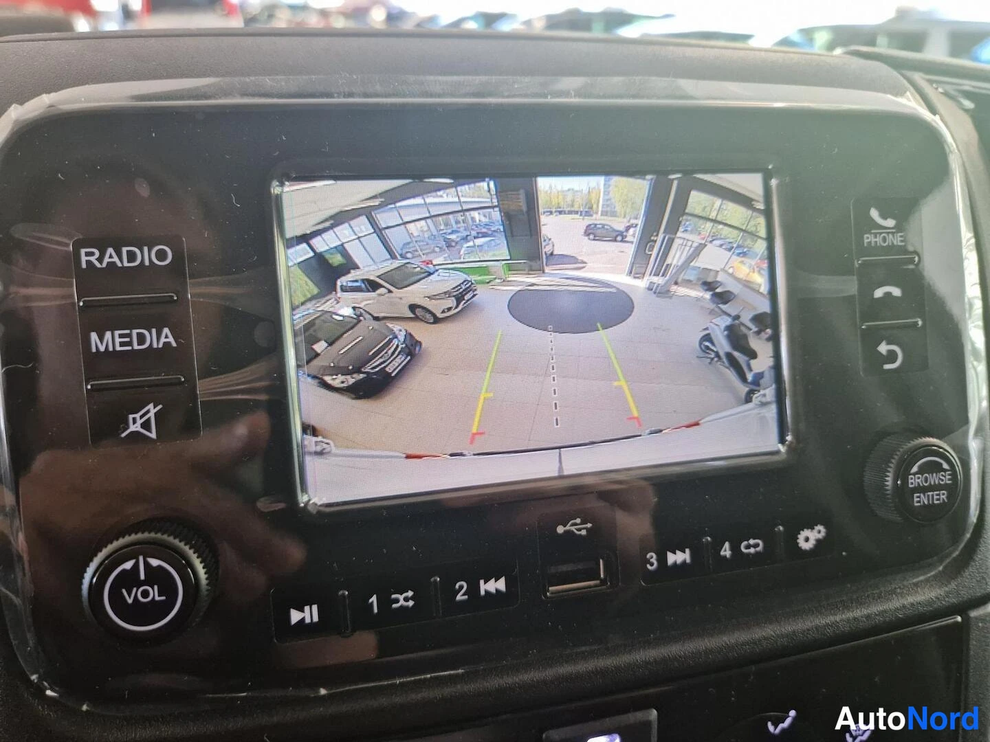 Yksityiskohta: Auton kojelaudan infotainment-yksikkö, jonka näytöllä on takakameran kuva ja ohjausviivat; Radio/Media-napit ja Vol-säädin näkyvissä.