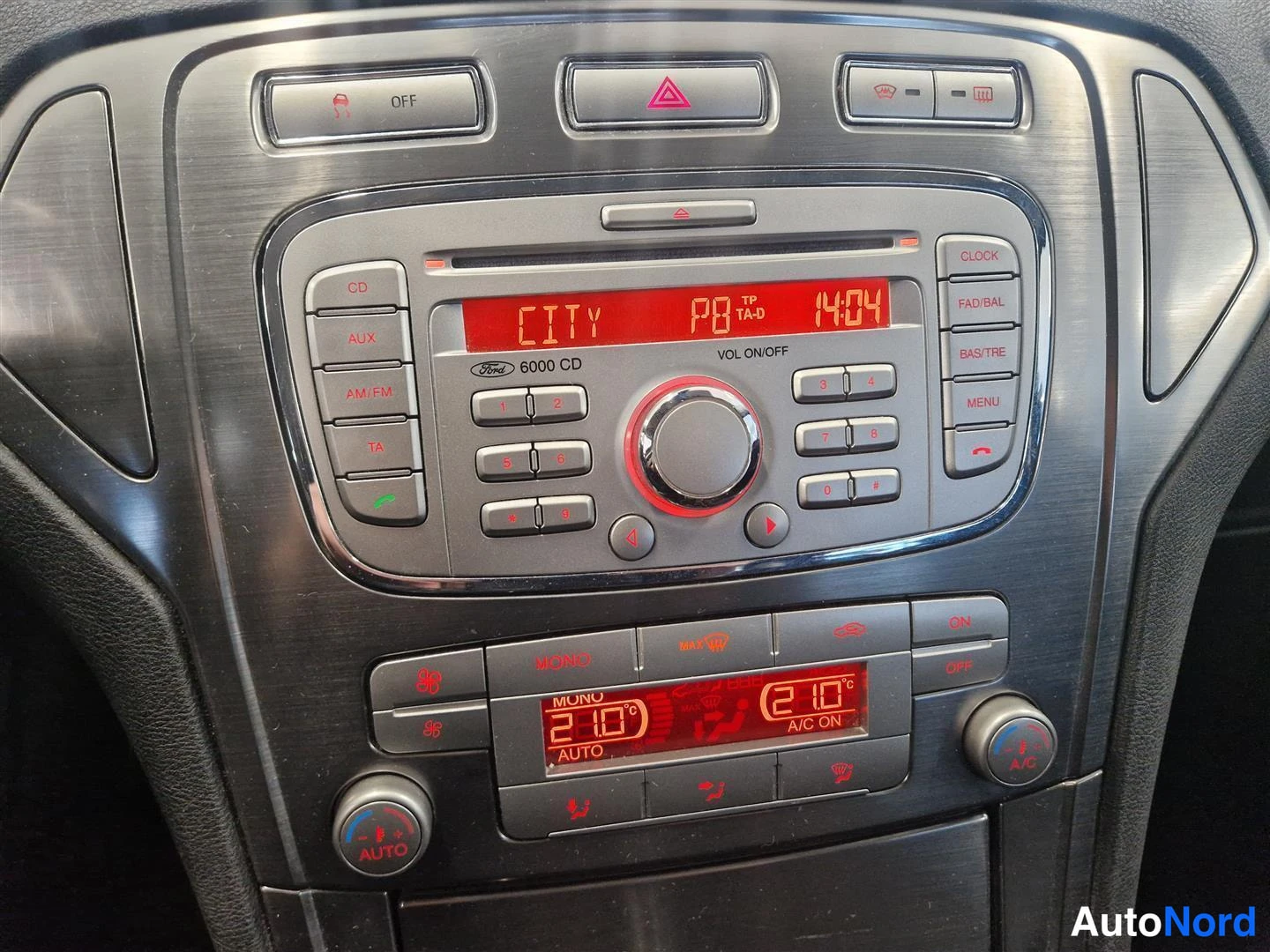 Etuviisto, nokka oikealle: Keskikonsoli: Ford 6000 CD -radio näytöllään CITY, TA-D, 14:04 ja automaattinen kaksialueinen ilmastointi (A/C ON, 21,0 °C, AUTO).